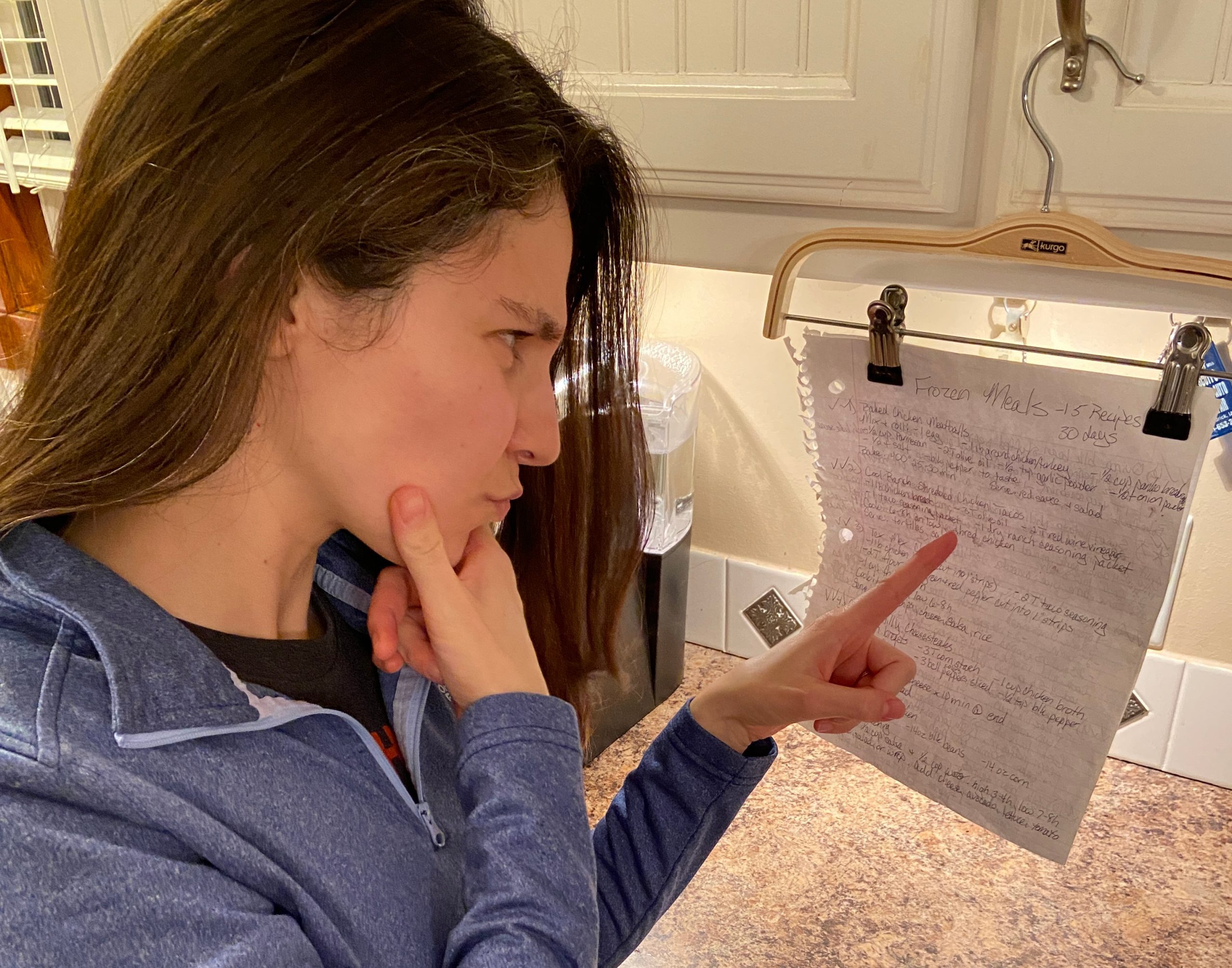 Woman looking at a recipe that is hanging from a kitchen cabinet on a pants hanger