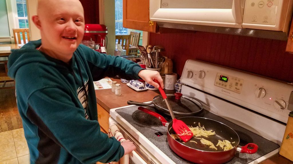 Sam, man with Down syndrome, stirring onions in a saute pan and smiling at the camera